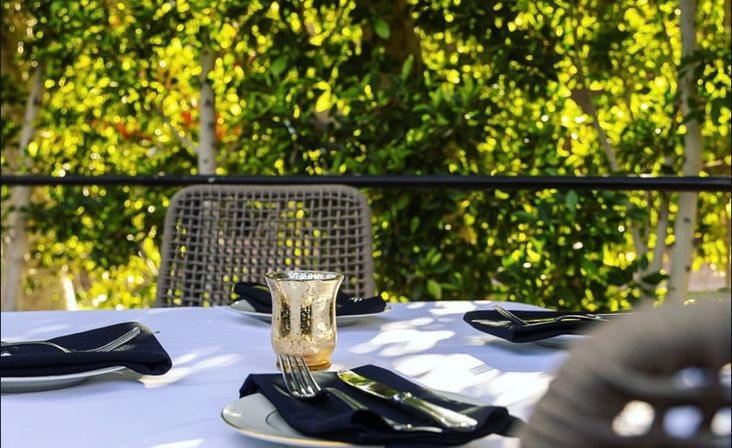 Sunlit outdoor patio dining table with white tablecloth, black napkins, silverware and a small gold votive glass, lush green foliage in the background.