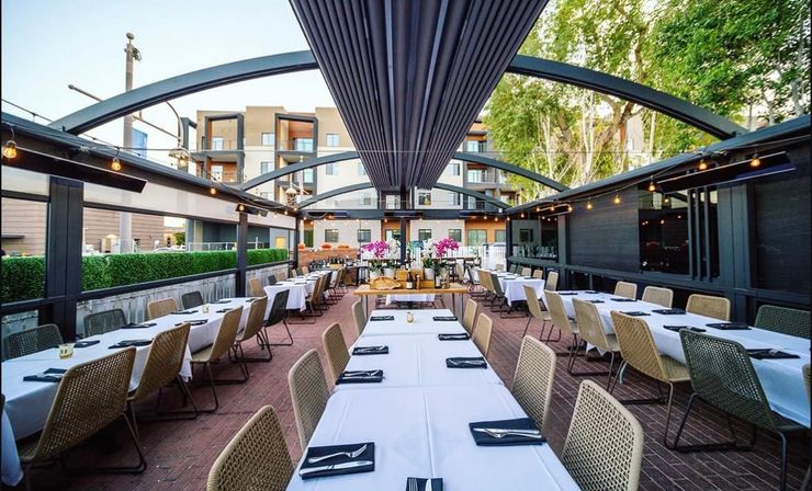 Covered outdoor restaurant patio with long white-tablecloth communal tables, woven chairs, hanging string lights and curved metal arches, set beside modern apartment buildings and trees for al fresco dining.