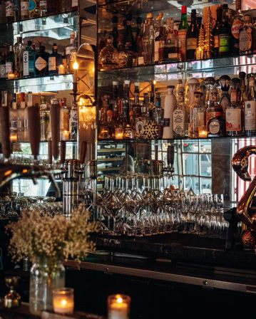 Cozy dimly lit cocktail bar back shelf with mirrored shelving filled with liquor bottles, stacked wine and martini glasses, beer taps, warm candlelight and a small vase of flowers.