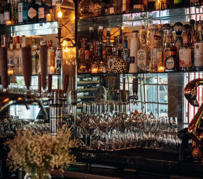 Cozy dimly lit cocktail bar back shelf with mirrored shelving filled with liquor bottles, stacked wine and martini glasses, beer taps, warm candlelight and a small vase of flowers.