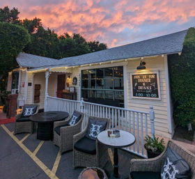 Cozy outdoor dining patio on a white porch with wicker chairs and round tables, string lights and a chalkboard sign for dinner and drinks under a pink sunset sky