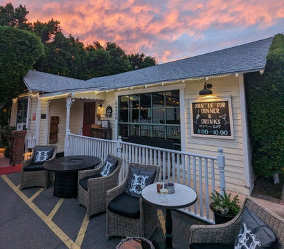 Cozy outdoor dining patio on a white porch with wicker chairs and round tables, string lights and a chalkboard sign for dinner and drinks under a pink sunset sky