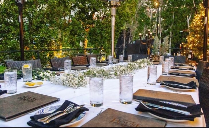 Al fresco patio dining table set for dinner with white tablecloth, tall water glasses (some with lemon), black napkins and silverware, baby's-breath centerpiece, wicker chairs and twinkling garden lights.