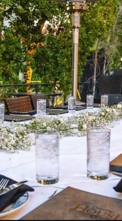Al fresco patio dining table set for dinner with white tablecloth, tall water glasses (some with lemon), black napkins and silverware, baby's-breath centerpiece, wicker chairs and twinkling garden lights.