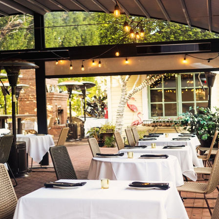 Al fresco restaurant patio with white-tablecloth tables, wicker chairs, hanging string lights and patio heaters under a covered pergola in a leafy garden setting