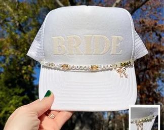 White trucker-style cap embroidered with 'BRIDE' in gold, adorned with a beaded bridal charm chain and tiny tassel, held outdoors against blue sky and autumn trees — bachelorette/bridal accessory.
