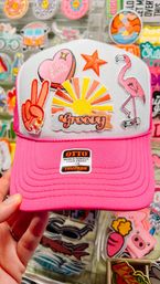Bright pink and white trucker hat with retro embroidered patches — yellow sunrise with ‘groovy’ text, pink flamingo on a skateboard, peace sign and ‘girl gang’ heart — shown against a colorful retail sticker display