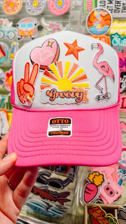 Bright pink and white trucker hat with retro embroidered patches — yellow sunrise with ‘groovy’ text, pink flamingo on a skateboard, peace sign and ‘girl gang’ heart — shown against a colorful retail sticker display