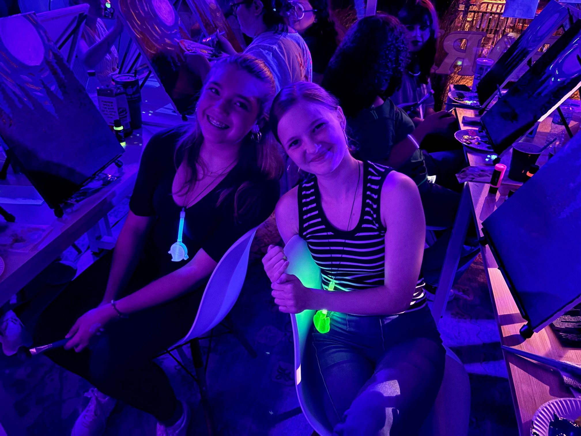 Two smiling young women at a neon paint-night workshop, seated at easels with paintbrushes in a dim indoor art studio bathed in purple and green glow-in-the-dark lighting.