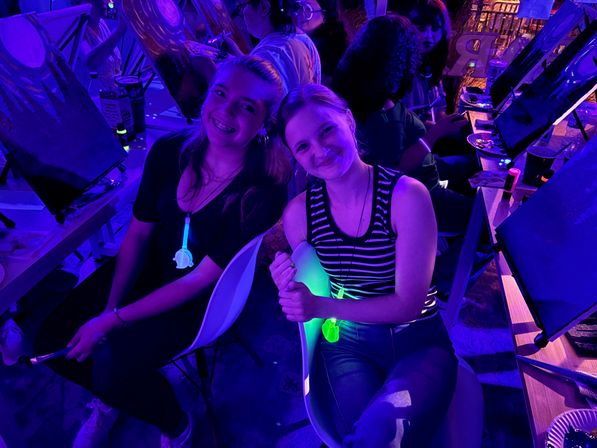 Two smiling young women at a neon paint-night workshop, seated at easels with paintbrushes in a dim indoor art studio bathed in purple and green glow-in-the-dark lighting.