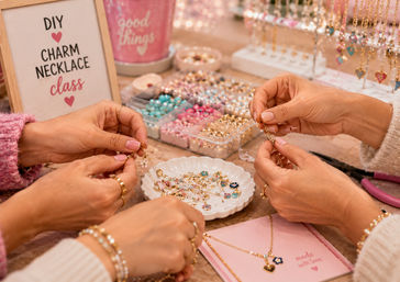 Hands assembling gold charm necklaces at a pink DIY charm necklace class workshop, with pastel beads, heart and flower charms, bead organizers and tools on the table