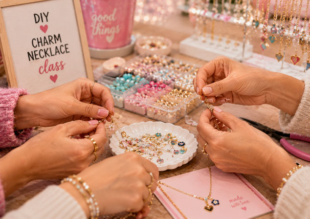 Hands assembling gold charm necklaces at a pink DIY charm necklace class workshop, with pastel beads, heart and flower charms, bead organizers and tools on the table
