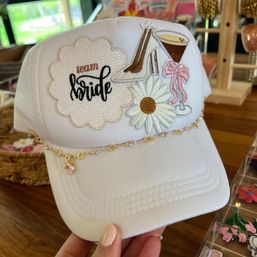 White trucker bridal hat with embroidered "team bride" patch, daisy, high heel and martini motifs, and a delicate gold chain across the brim — bachelorette party wedding accessory.