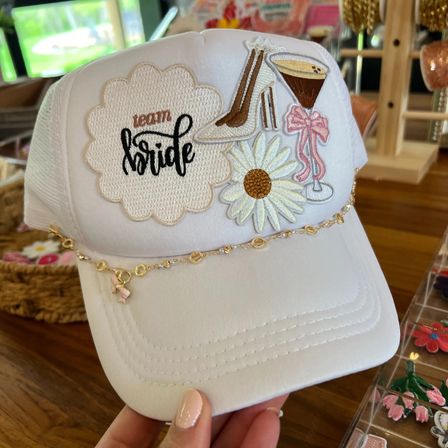 White trucker bridal hat with embroidered "team bride" patch, daisy, high heel and martini motifs, and a delicate gold chain across the brim — bachelorette party wedding accessory.