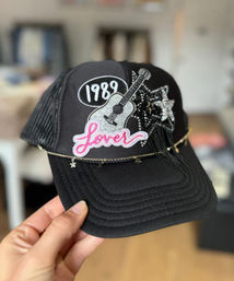 Playful black trucker hat with embroidered 1989 patch, pink Lover patch, glitter guitar and star patches, and a delicate star chain draped across the brim, handheld against a blurred indoor background