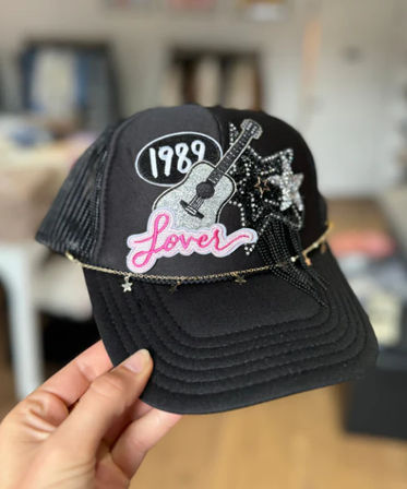 Playful black trucker hat with embroidered 1989 patch, pink Lover patch, glitter guitar and star patches, and a delicate star chain draped across the brim, handheld against a blurred indoor background