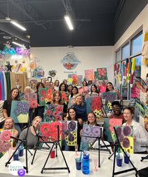 Smiling adults in a bright art studio in Summerville, SC holding colorful hand-painted floral canvases during a guided paint-night group class