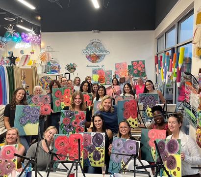 Smiling adults in a bright art studio in Summerville, SC holding colorful hand-painted floral canvases during a guided paint-night group class