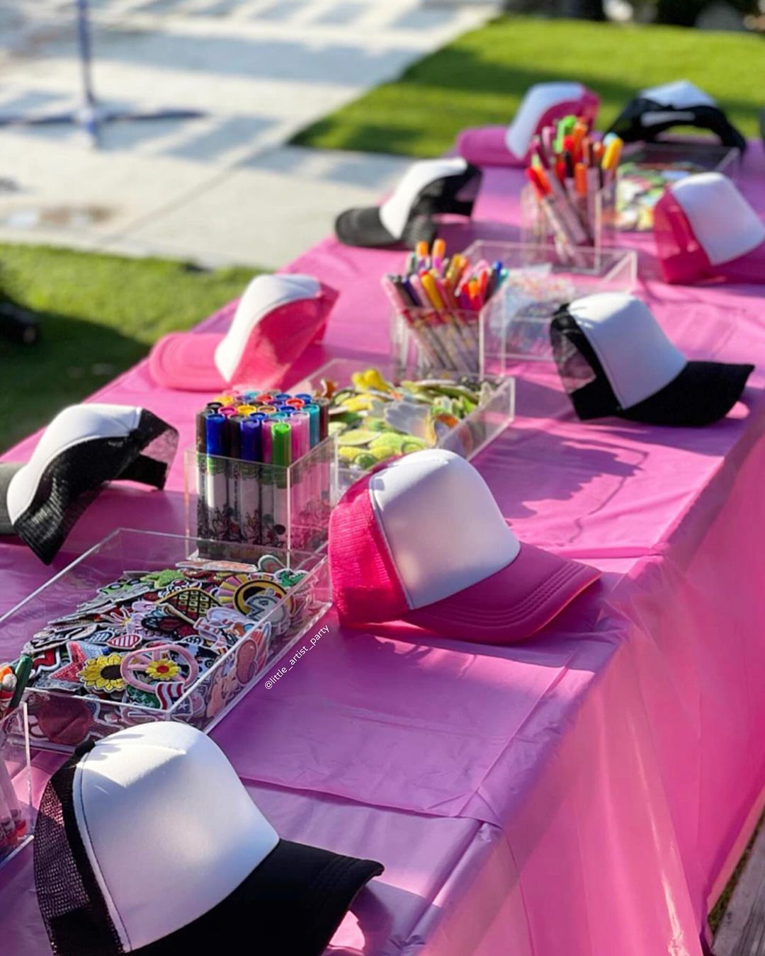 Outdoor pink craft table set up for a DIY hat-decorating party with blank white-front trucker caps, colorful markers, and trays of stickers and embellishments.