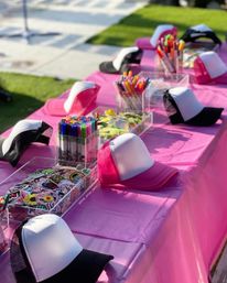 Outdoor pink craft table set up for a DIY hat-decorating party with blank white-front trucker caps, colorful markers, and trays of stickers and embellishments.