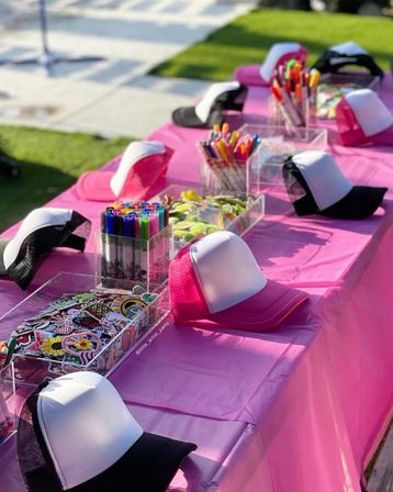 Outdoor pink craft table set up for a DIY hat-decorating party with blank white-front trucker caps, colorful markers, and trays of stickers and embellishments.