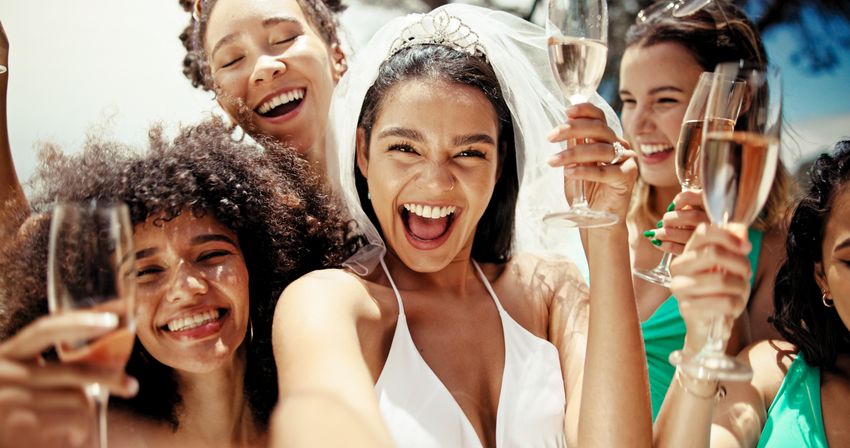 Bride wearing a veil and tiara laughing with friends as they toast champagne flutes in a sunny outdoor bachelorette party selfie.