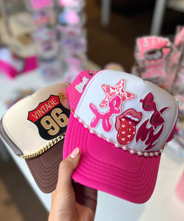 Hand holding two trendy trucker caps in a boutique: a hot pink-and-white embellished hat with pearl trim and playful patches (starfish, balloon dog, lips, sunglasses) and a brown-beige cap with a gold-chain detail and "Vintage 96" patch, blurred pink shop display in background.