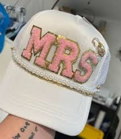 White trucker hat bridal accessory featuring large pink glitter 'MRS' letters outlined in gold, pearl and gold-chain trim on the brim and a small ring charm, bachelorette/wedding hat.