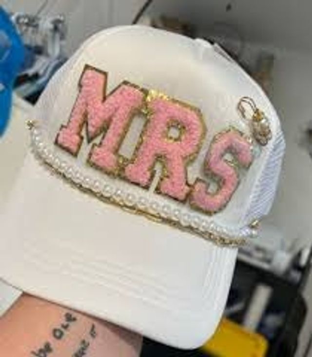 White trucker hat bridal accessory featuring large pink glitter 'MRS' letters outlined in gold, pearl and gold-chain trim on the brim and a small ring charm, bachelorette/wedding hat.
