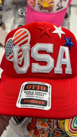 Patriotic red foam-front trucker hat embroidered with bold white "USA", stitched stars and a heart-shaped American flag patch, retail sticker on the brim, displayed on a shelf.