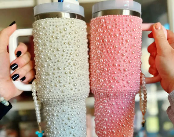 Two pearl-covered insulated travel tumblers — one white and one pink — with handles and mouse-ear straw charms, held side-by-side by two hands against a blurred shelf background.
