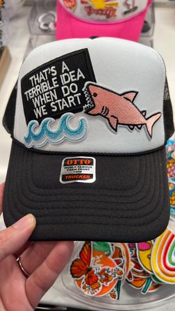 Black-and-white foam-front trucker hat with embroidered pink shark, blue waves, and a patch reading "THAT'S A TERRIBLE IDEA WHEN DO WE START", held by a hand.