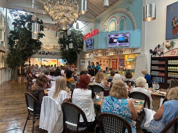 Busy coastal‑themed cooking class in a bright indoor kitchen: long communal tables of attendees, three instructors at a demonstration counter beneath a pastel mural and neon sign, oversized pan chandelier, leafy indoor trees and rooster décor.