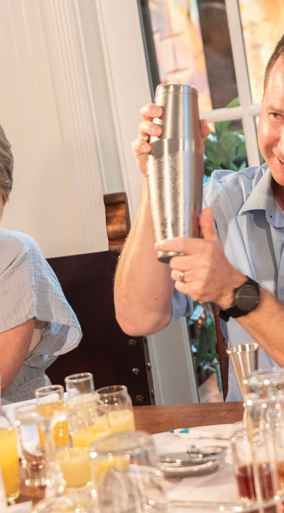 Two people playfully shaking stainless steel cocktail shakers at a dining table crowded with glasses and orange juice in a bright indoor setting