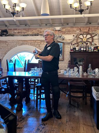 Person demonstrating bartending technique in a nautical-themed brick bar interior with a ship’s wheel, coiled rope, wooden table, chandeliers and bottles on a cabinet.