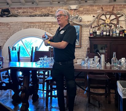 Person demonstrating bartending technique in a nautical-themed brick bar interior with a ship’s wheel, coiled rope, wooden table, chandeliers and bottles on a cabinet.