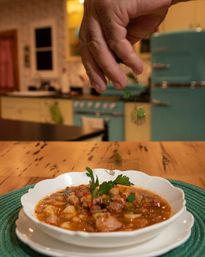 Hand sprinkling fresh parsley over a steaming bowl of homemade soup on a wooden table in a cozy retro teal kitchen.