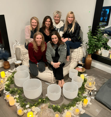 Six women seated on floor cushions in a cozy living room around a circle of crystal singing bowls, LED candles and greenery for a sound bath meditation gathering.
