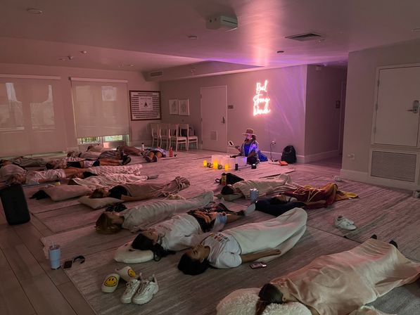 Group meditation/sound-bath in a dim wellness studio — participants lying on mats under blankets around candles with a cozy pink neon glow and relaxed, cozy vibe.