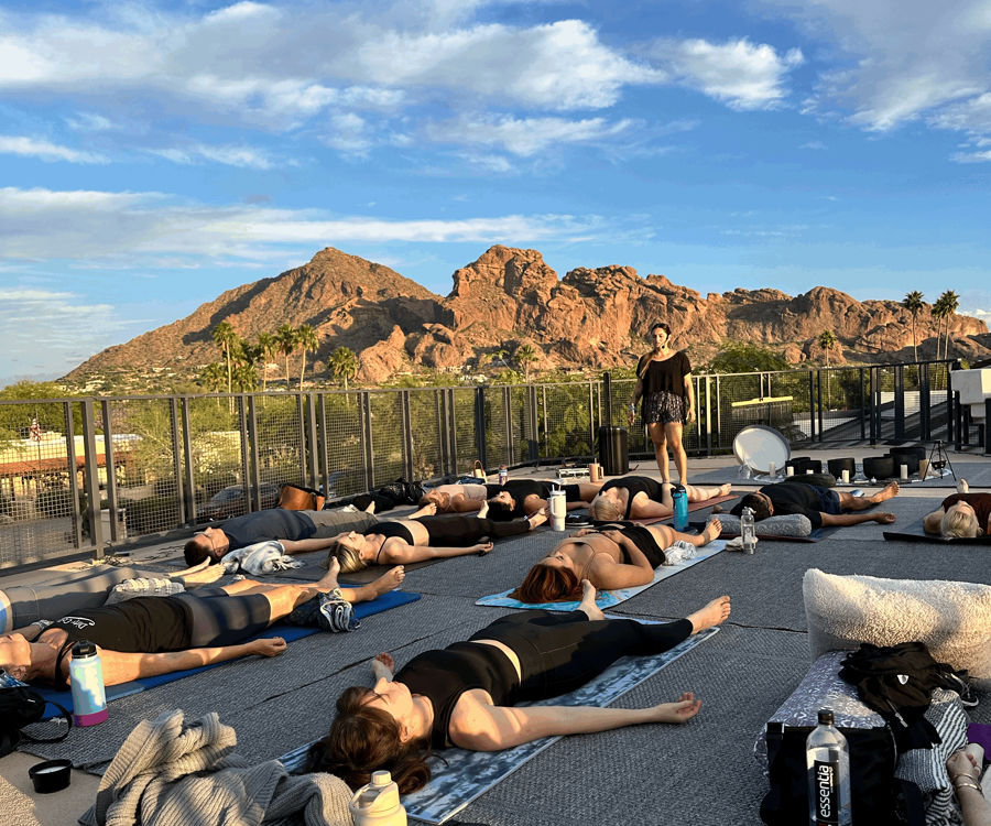 Rooftop sunset yoga class in a desert city—students lying on mats in Savasana while an instructor stands, red rock mountains and palm trees under a bright blue sky.