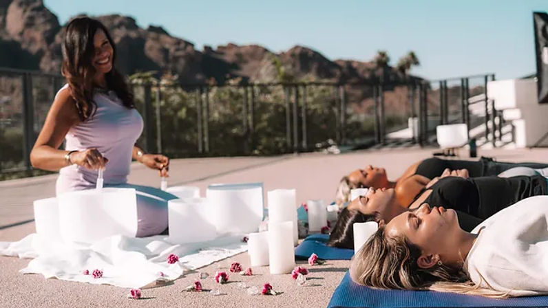 Rooftop sound bath session with a facilitator playing crystal singing bowls while participants lie on mats among candles and rose petals with a sunlit hillside backdrop.