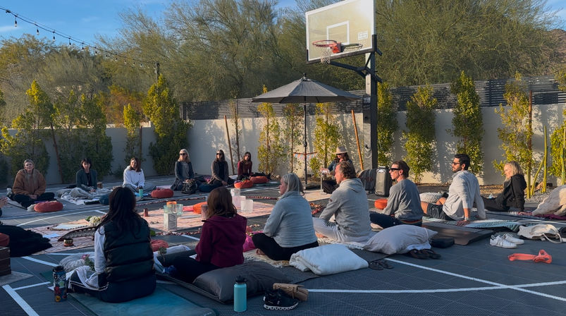 Outdoor meditation circle on a residential basketball court at golden hour — participants seated on cushions and mats around blankets and candles, with desert trees, string lights and a basketball hoop in the background.