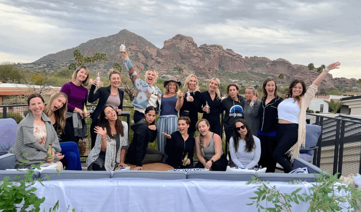 Rooftop group of women laughing and raising drinks on a patio with Arizona desert rock formations and palm trees in the background