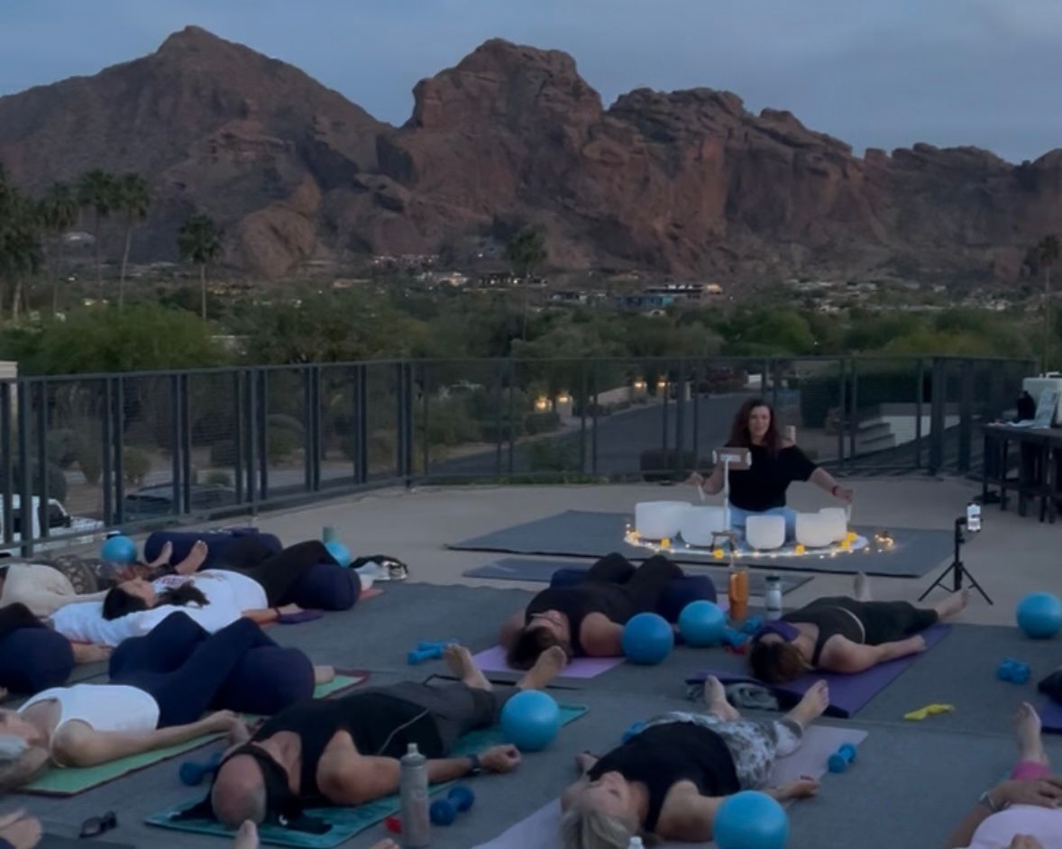 Rooftop sunset sound-bath yoga class with participants lying in savasana, blue exercise balls and crystal singing bowls, desert red‑rock mountains in the background.