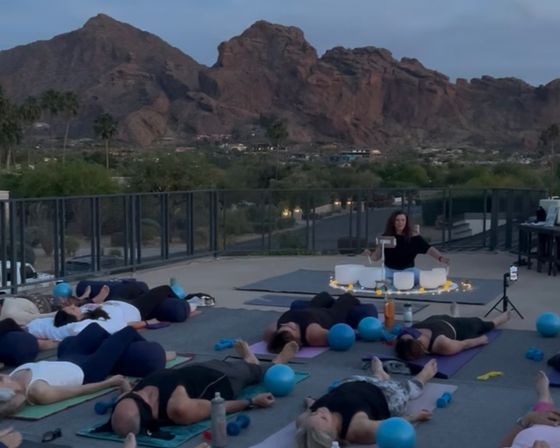 Rooftop sunset sound-bath yoga class with participants lying in savasana, blue exercise balls and crystal singing bowls, desert red‑rock mountains in the background.