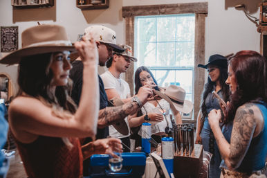 Group of shoppers trying on and shaping cowboy hats in a rustic western hat shop, staff using a small torch to mold a hat brim by a sunlit window.