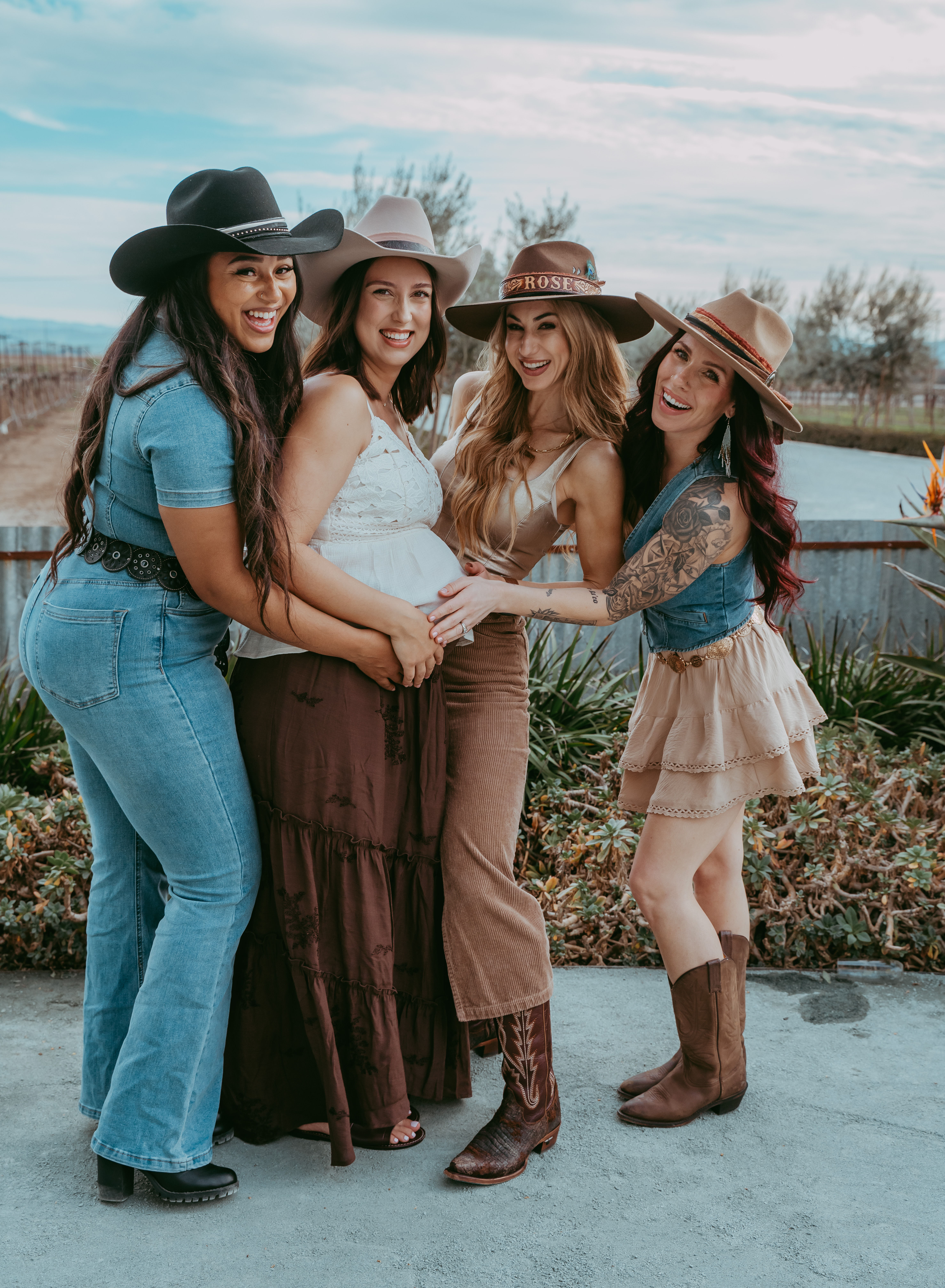 Four smiling women in cowgirl hats and western outfits, one pregnant, posing and touching her baby bump outdoors by a rustic fence and pond