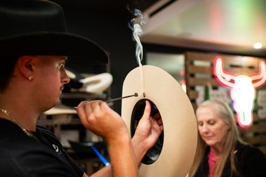Artisan hat maker shaping a beige felt cowboy hat with a steaming tool in a busy hat workshop, neon bull sign and a customer blurred in the background.