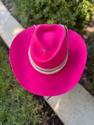Top-down view of a vibrant hot-pink felt cowboy hat with a beige decorative band resting on grass beside a concrete edge.