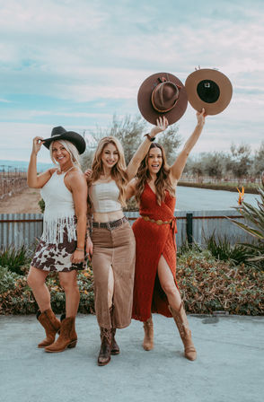 Three smiling women in boho country outfits and cowgirl boots pose in front of vineyard rows and a corrugated metal fence, two holding wide-brim hats aloft against a blue sky.